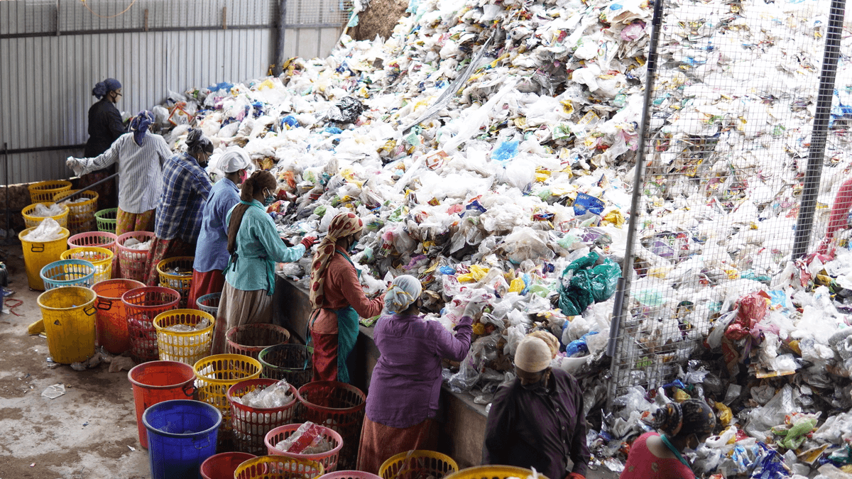 Workers sorting through a large pile of garbage in a recycling facility. They are wearing protective gear such as gloves and masks, and are placing sorted materials into colorful plastic bins. The area is enclosed with metal fencing.